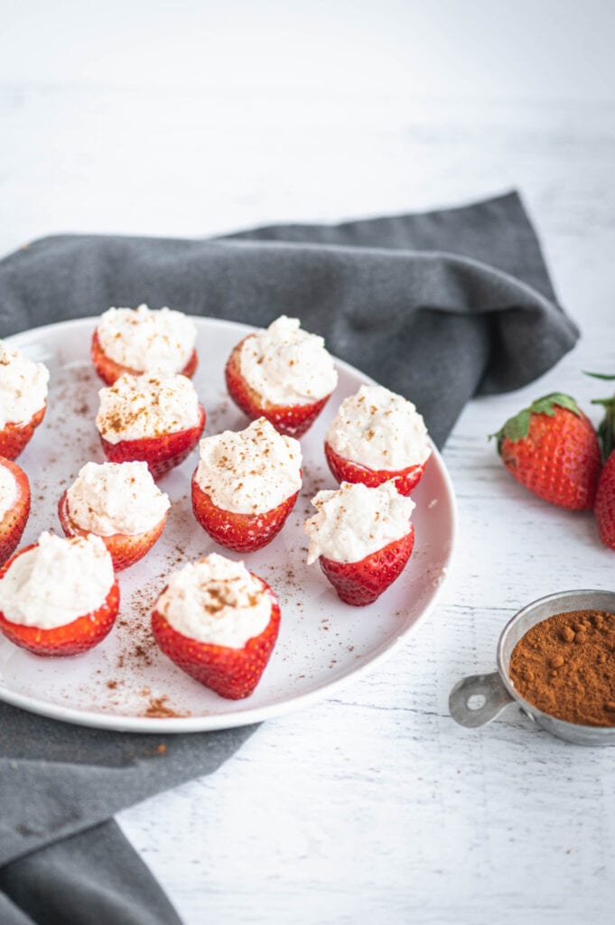 High angle photo of cheesecake stuffed strawberries standing on end on a white plate. There is a bowl of cinnamon and whole strawberries off to the side with a gray towel.