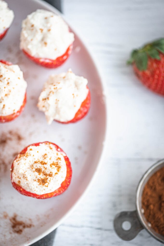 Overhead photo of Cheesecake Stuffed Strawberries with homemade whipped ricotta on a round white plate on a white wooden background. Off to the side is a bowl of cinnamon and a whole strawberry.