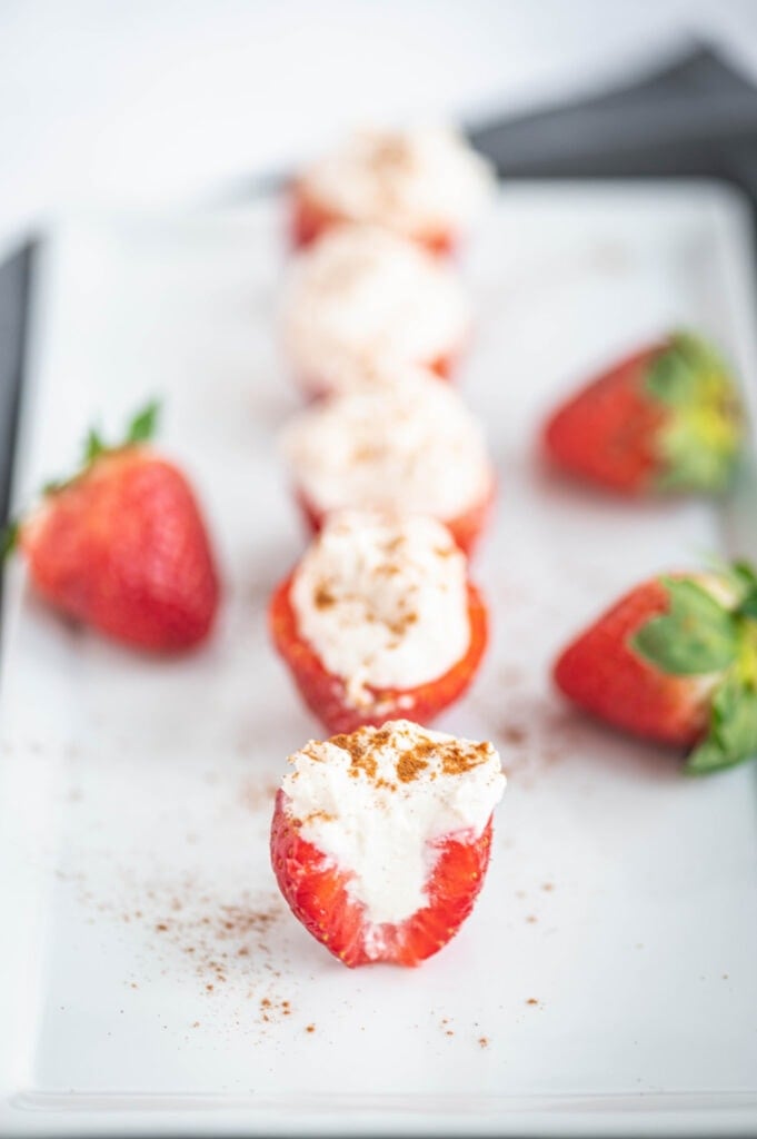 Side photo of a cross section of a cheesecake stuffed strawberry topped with cinnamon. In the background, out of focus, are more stuffed strawberries and whole strawberries on the serving dish.