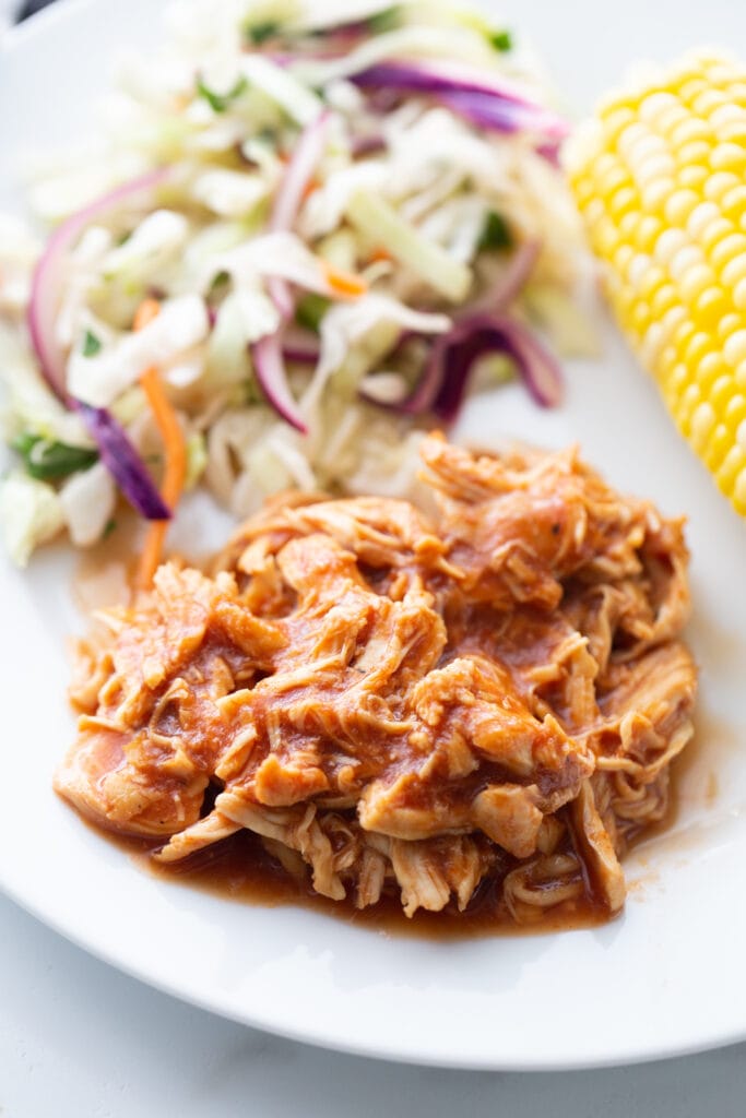 Close up photo of a white plate filled with pulled BBQ chicken. Cilantro lime slaw and corn on the cob are also on the plate, in the background, slightly out of focus.