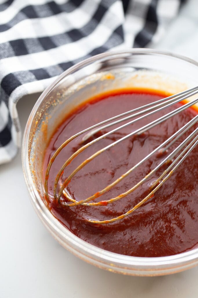 Glass bowl full of homemade BBQ sauce just whisked together, with a navy and white checkered towel in the background.