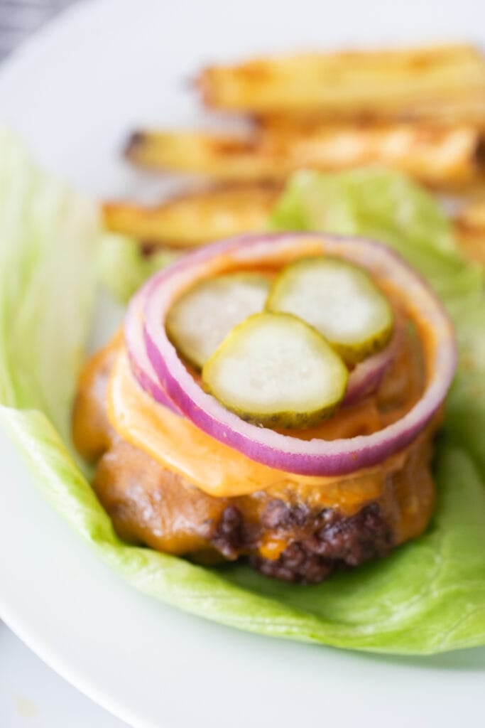 Close of photo of a bunless burger topped with cheddar cheese, burger sauce, red onion, and pickles, on a lettuce leaf. In the background, there are french fries on the plate also, out of focus.