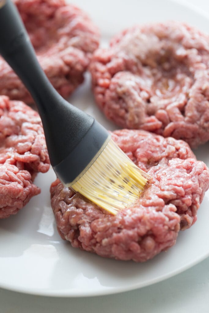 Raw burger patties on a white plate being brushed with olive oil