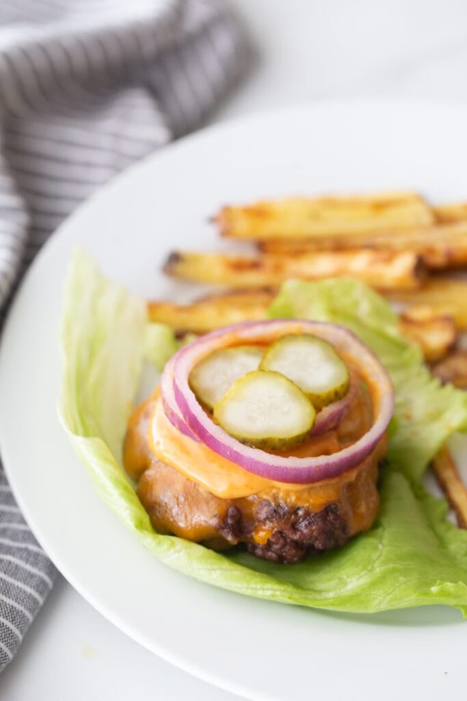A bunless burger topped with cheddar cheese, burger sauce, red onion, and pickles, on a lettuce leaf. There are french fries on the plate also, out of focus.