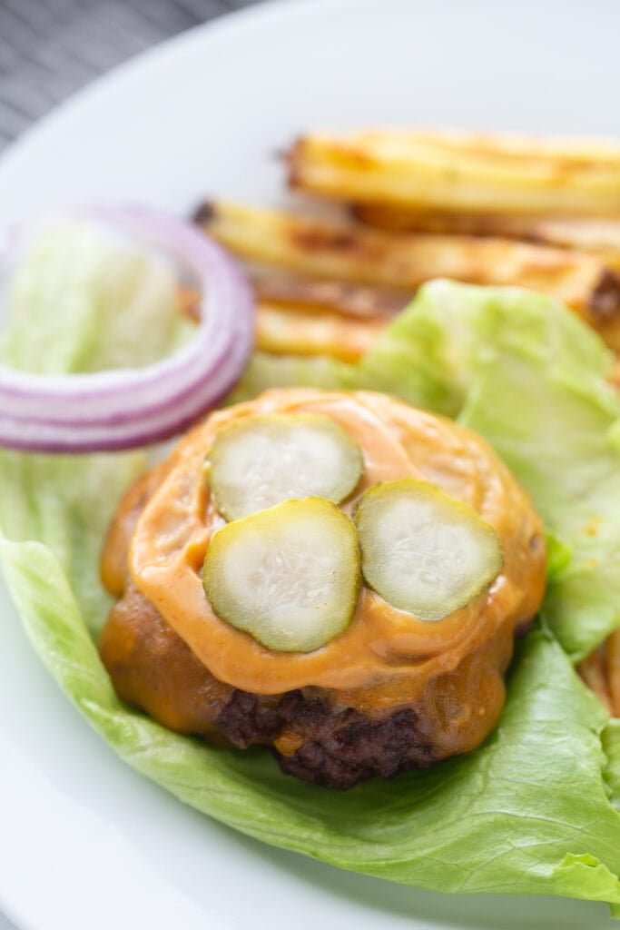 The best bunless burger recipe topped with cheddar cheese, burger sauce,, and pickles, on a lettuce leaf. There are red onions and french fries on the plate also, out of focus.
