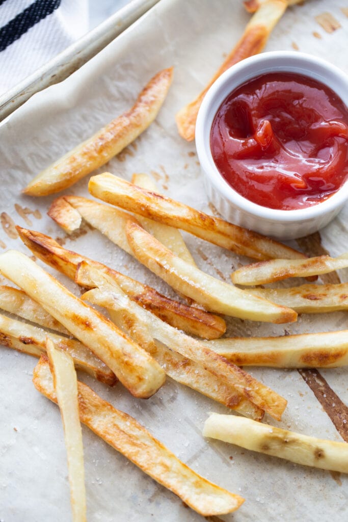 Overhead photo of golden brown homemade baked French Fries on a parchment lined sheet pan. There is a white ramekin of ketchup on the corner of the pan. 