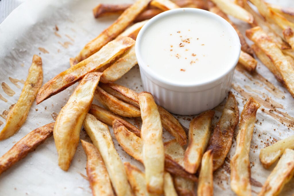 Parchment lined baking sheet filled with crispy crab seasoned French fries and a ramekin filled with homemade cheese sauce.