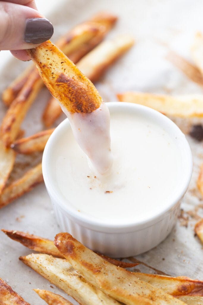 Close up photo of a woman's hand holding a homemade seasoned French fry dipping into homemade cheese sauce in a white ramekin. The ramekin sits on a parchment lined baking sheet filled with crispy French fries.
