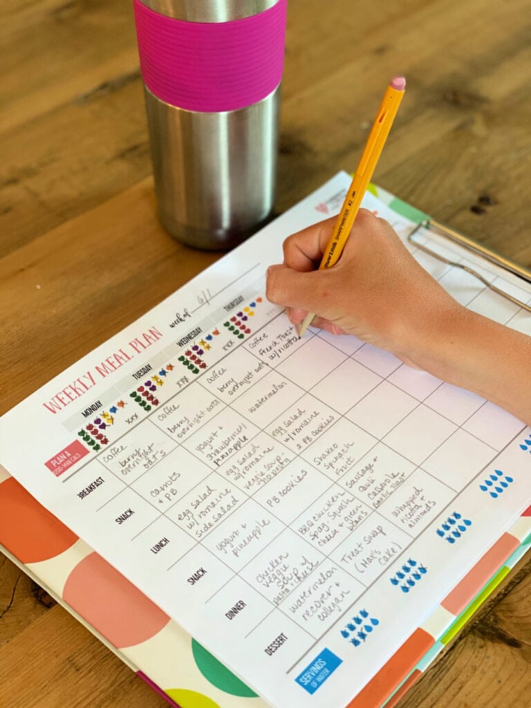 A hand holding a pencil and writing in a 21 Day Fix meal planner tracking sheet on a colorful clip board on a wooden table. There is a metal water bottle with a pink grip at the top of the photo.