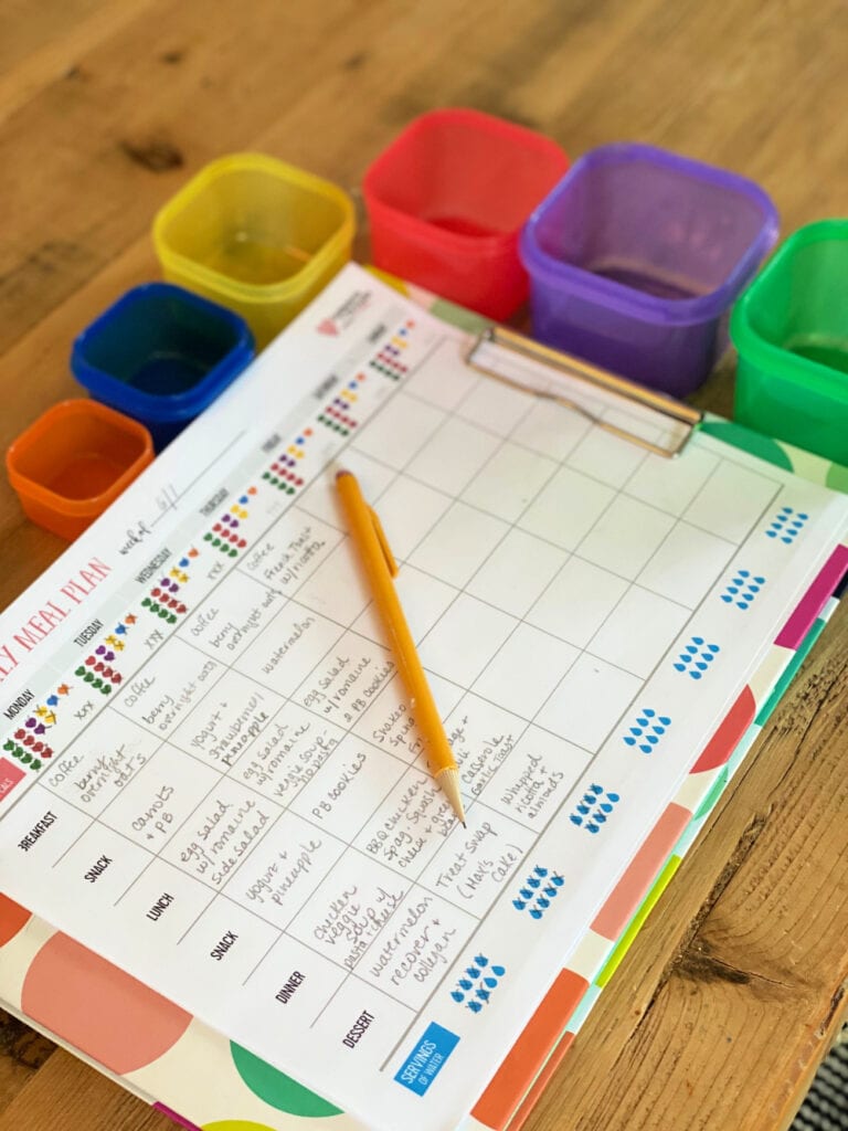 Color coded paper meal tracking sheet for the 21 Day Fix on a colorful clip board with a pencil on top. Next to the clip board are the colored containers for tracking food on the 21 Day Fix.