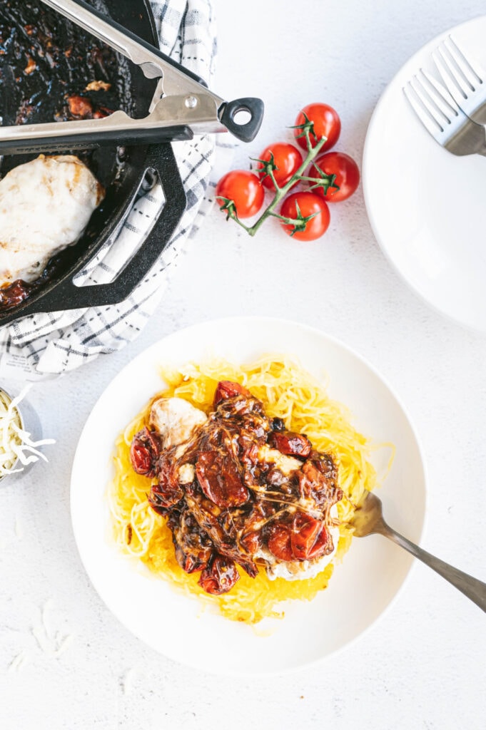 Overhead photo of a nice portion of spaghetti squash topped with caprese chicken. The skillet where the chicken was cooked is off to the side of the photo with more chicken, ready to be served. There are vine ripe tomatoes off to the side as well. 