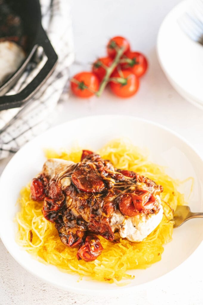 Close up photo of one skillet caprese chicken. Melted cheese, balsamic reduction, and juicy tomatoes top a chicken breast over spaghetti squash in a white bowl. In the background, out of focus, are more tomatoes, extra plates, and the cast iron skillet used to cook the chicken.