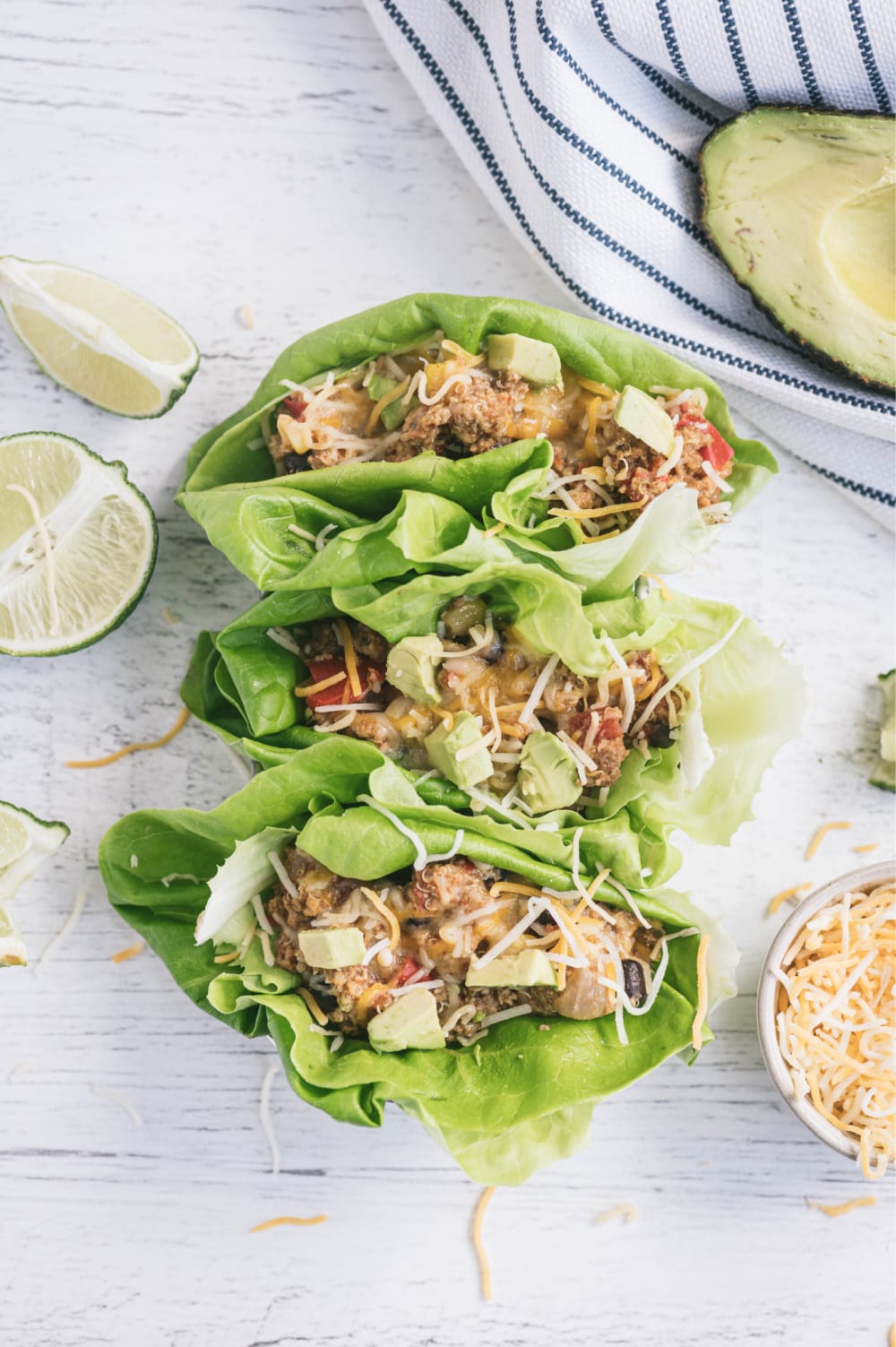 Overhead photo with lettuce wraps filled with Mexican crock pot taco casserole on a white surface. Surrounding the wraps are a white and blue striped towel, half an avocado, sliced limes, and a ramekin full of shredded cheese.