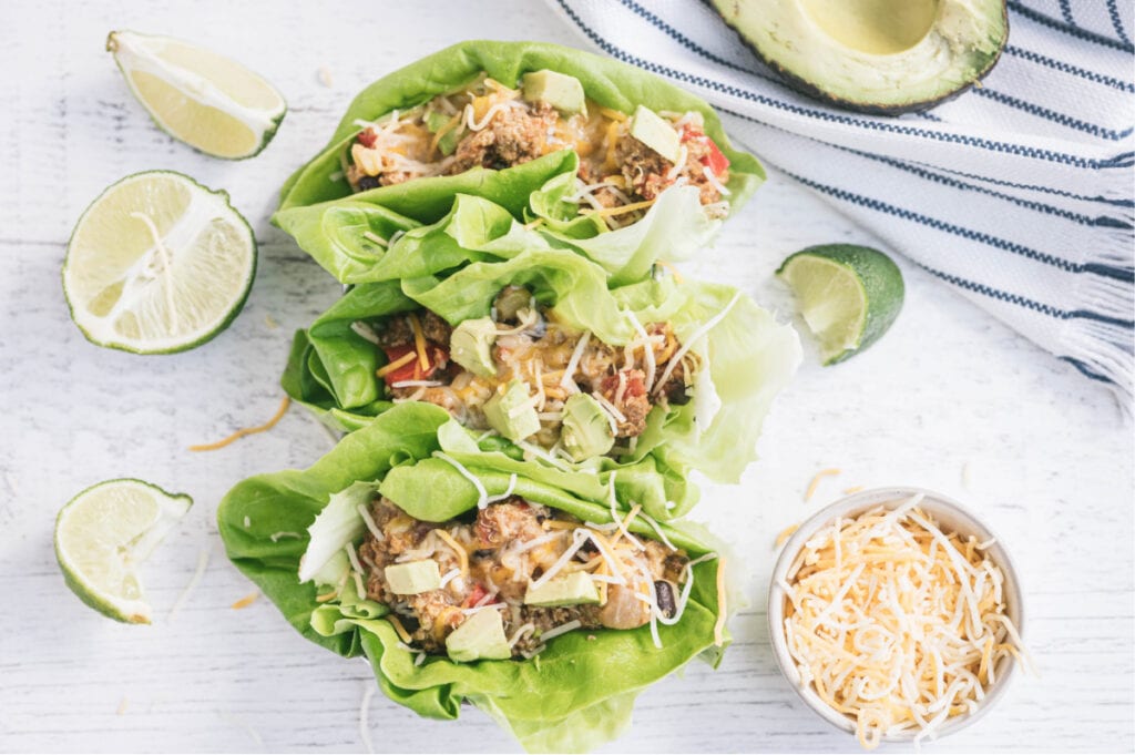 Three lettuce wraps filled with crock pot taco casserole on a white surface. Surrounding the wraps are a white and blue striped towel, half an avocado, sliced limes, and a ramekin full of shredded cheese.