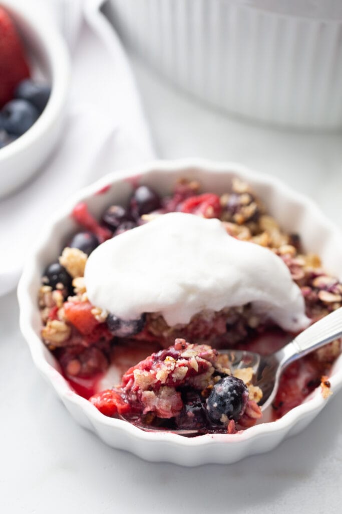 Individual berry crisp topped with coconut cream. There is a spoon with a bit of crisp, ready to eat.