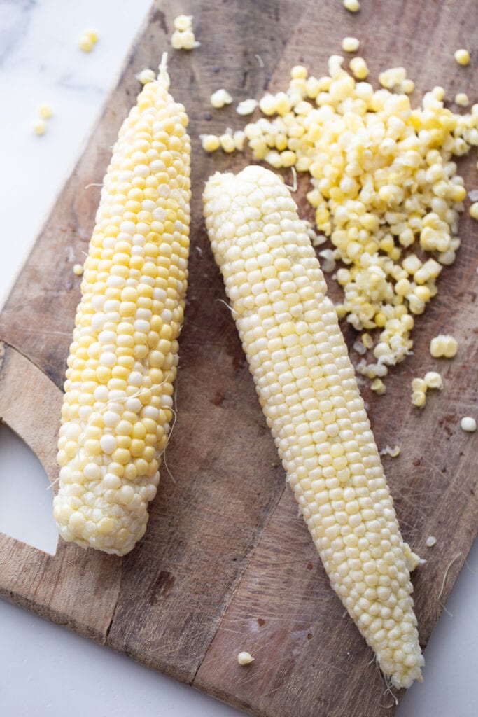 two ears of corn with freshly shucked corn kernels sitting on a wooden cutting board. 
