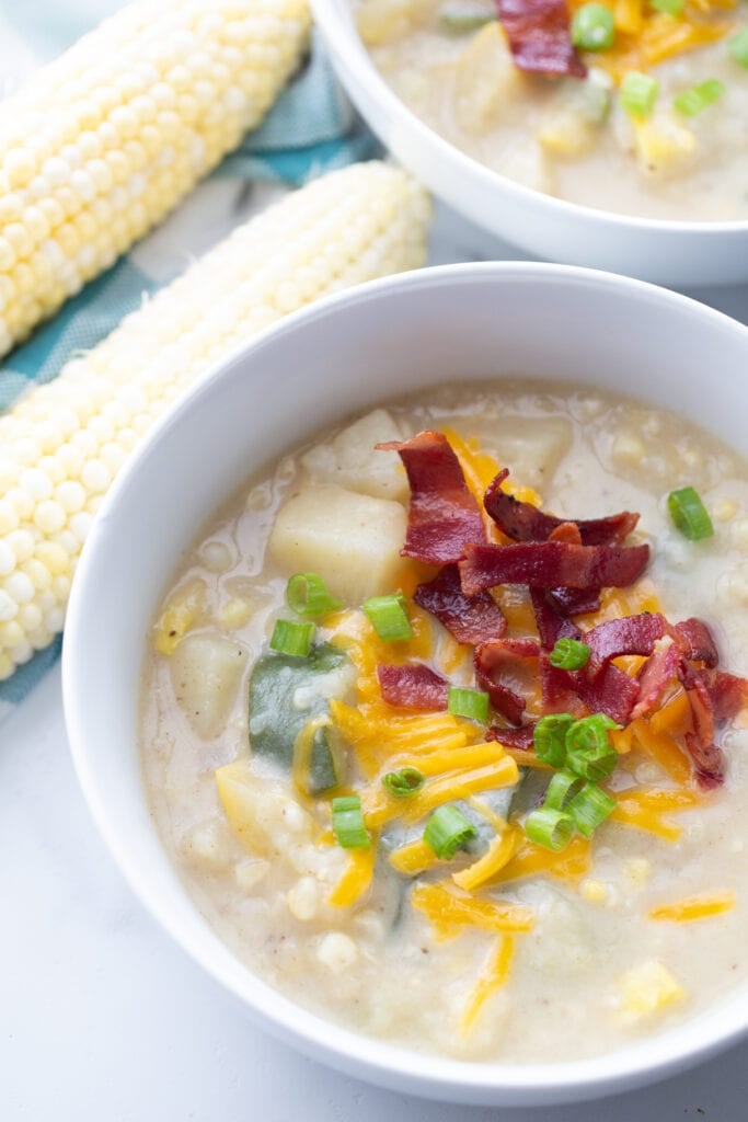 A white bowl filled with fresh corn chowder and topped with cheddar cheese and turkey bacon. Two ears of corn and a second bowl of chowder are seen in the background