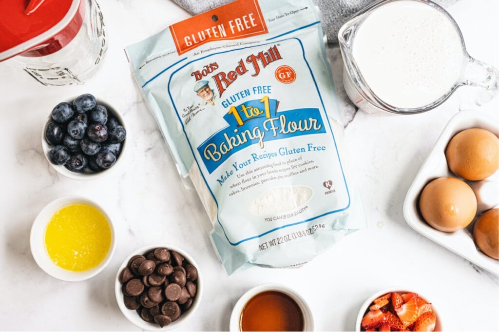 A bag of Bob's Red Mill Gluten Free 1 to 1 Baking Flour is on it's side and centered in the photo. All around the bag of flour are ingredients to make Gluten Free Pancakes from scratch.