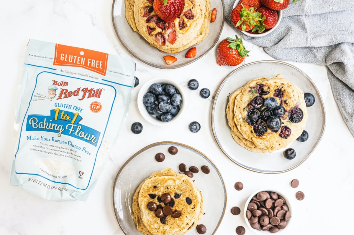 Overhead photo showing plates of pancakes topped with fruit or chocolate chips. A bag of Bob's Red Mill Gluten Free 1 to 1 Baking Flour is off to the side showing the wonderful base for cooking the Gluten Free pancakes.