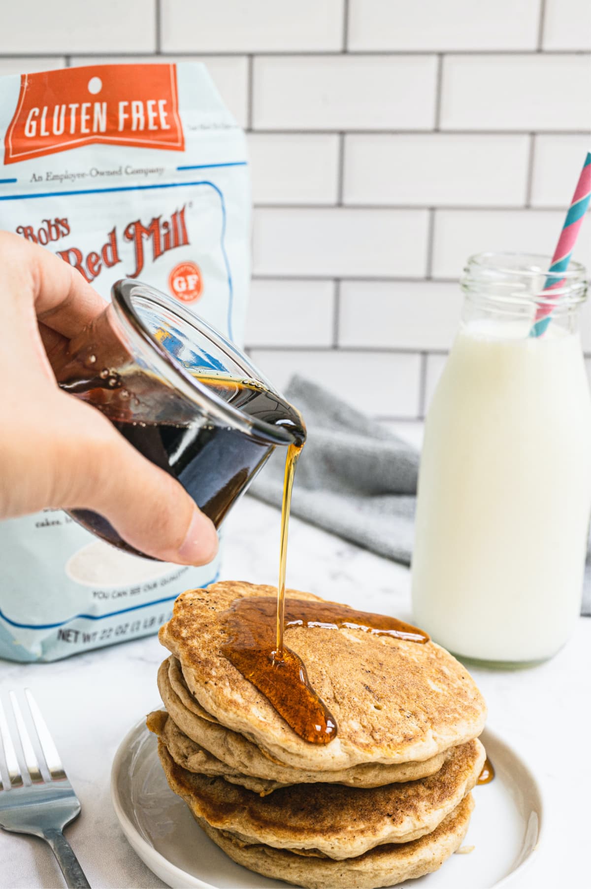 A person's hand pouring a glass container of syrup onto a stack of pancakes with a glass bottle of milk in the background.
