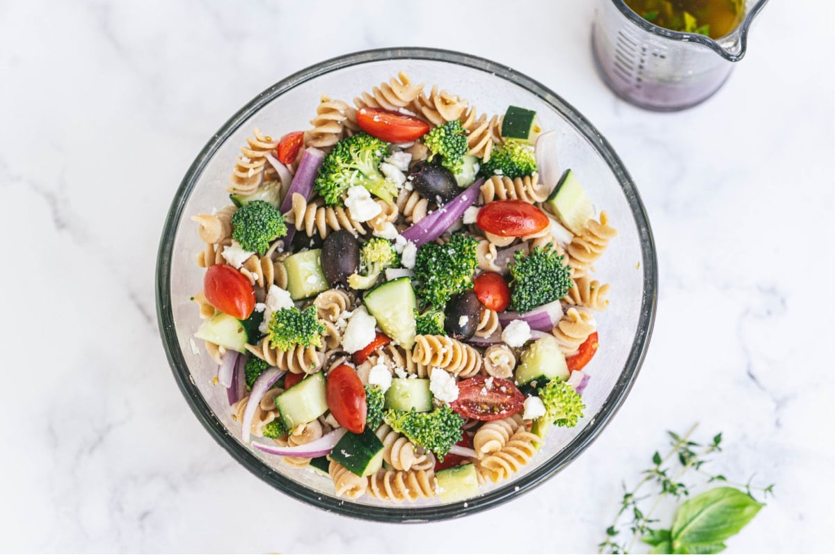 A large bowl of pasta salad with tomatoes, cucumbers, broccoli, red onions, feta cheese, olives, and pasta. There is dressing in a small pitcher off to the side.