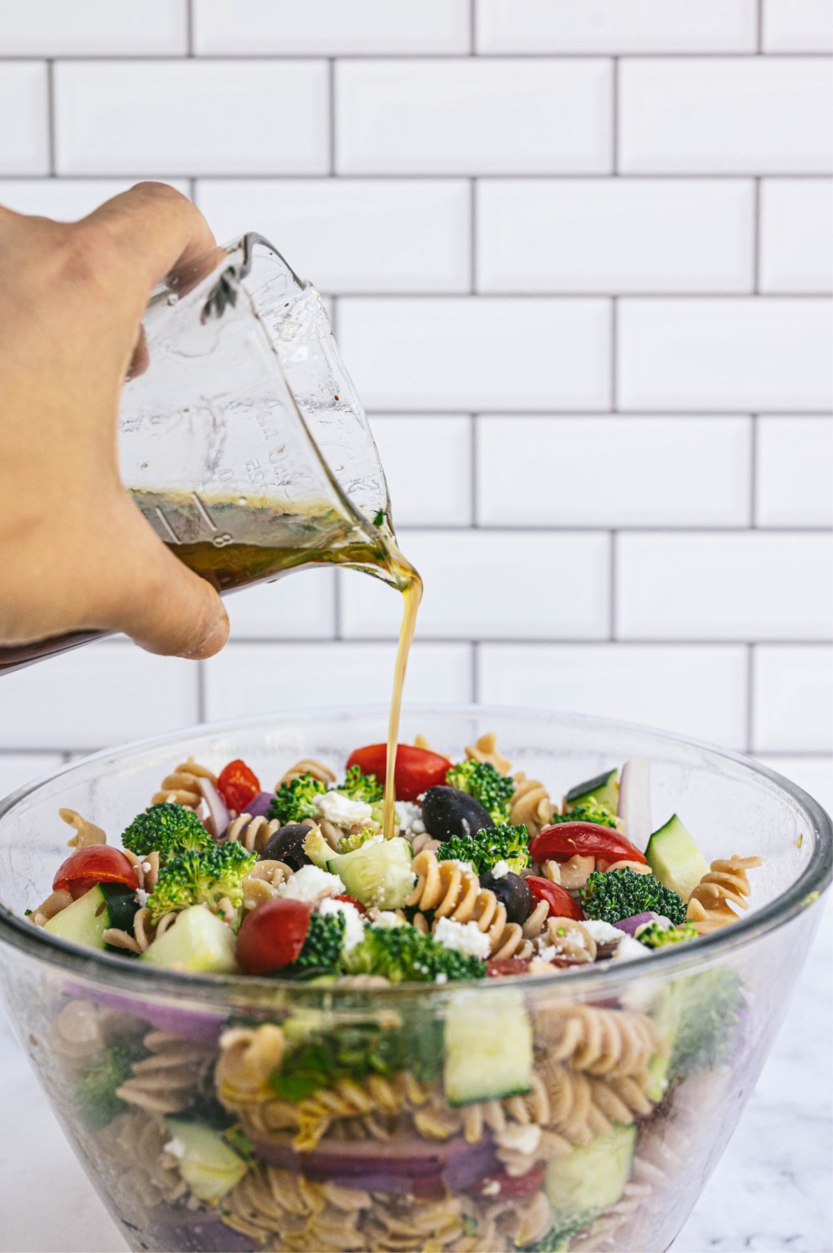 A hand pouring dressing from a glass container into a bowl bowl of Greek pasta salad with white subway tiles in the background.