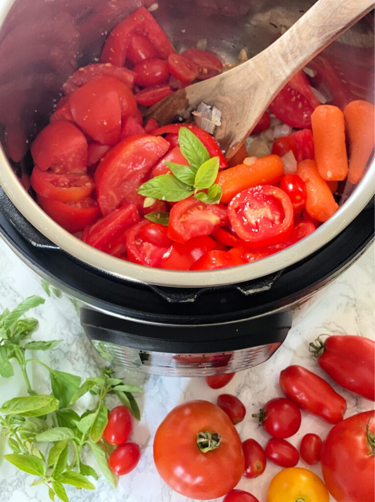 Roughly chopped garden tomatoes, basil, and baby carrots in the Instant Pot, ready for homemade tomato sauce. Basil and whole tomatoes are in front of the pot as well.
