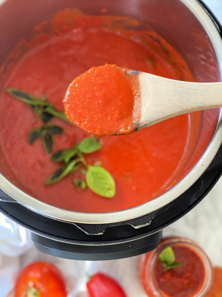 Overhead photo of tomato sauce on a wooden spoon. In the background is the rest of the pot of sauce and a smaller jar full of homemade sauce.
