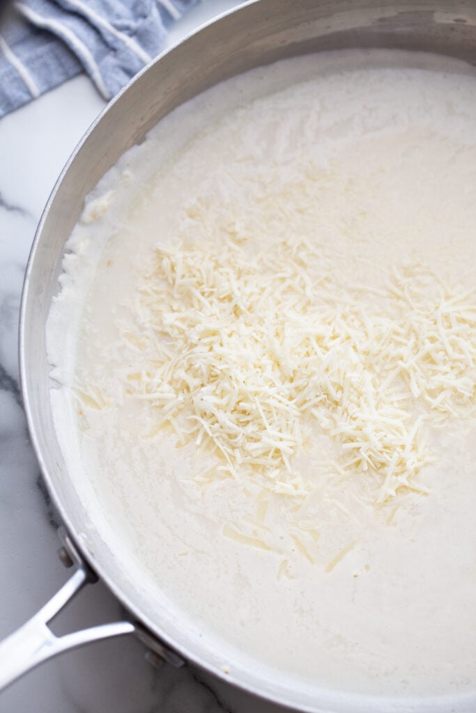 Shredded cheese beginning to melt in a thickened Alfredo sauce with milk in a large, shallow metal pan.
