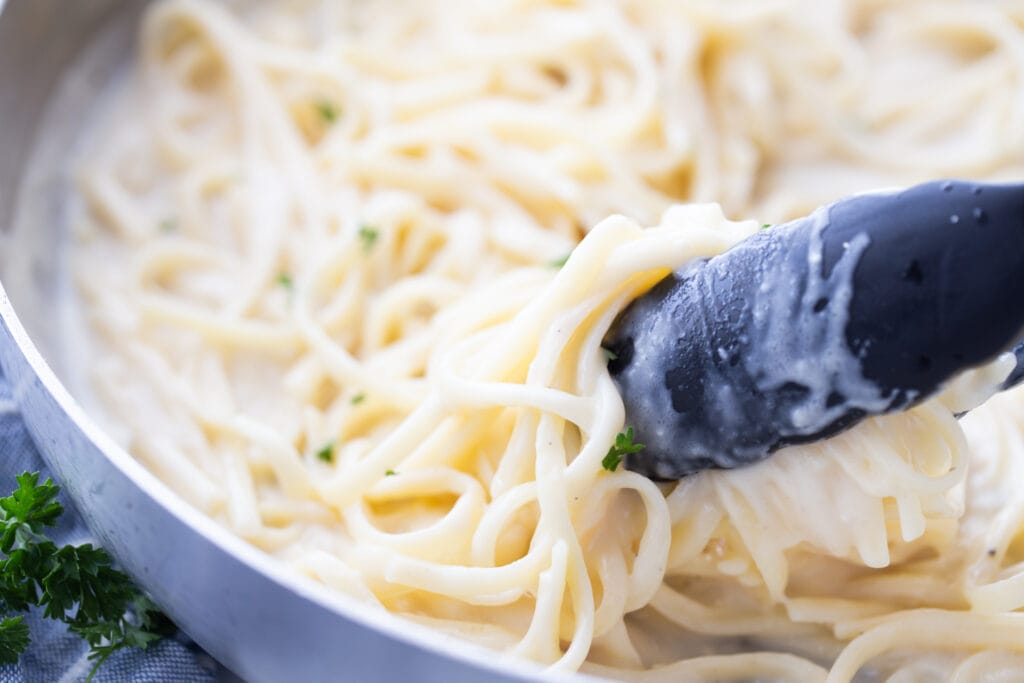 Close up photo of a shallow pan of fettuccine tossed with alfredo sauce. There are black tongs grabbing a portion of pasta out of the pan.