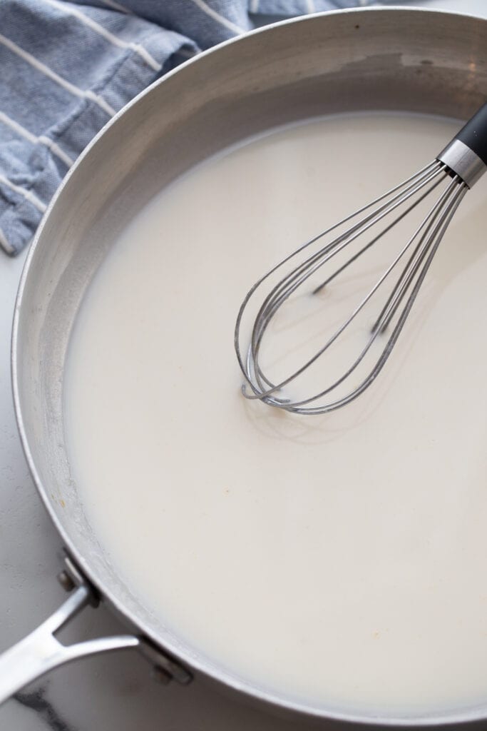 Black handled metal whisk resting on the side of a shallow pan with a white sauce inside.