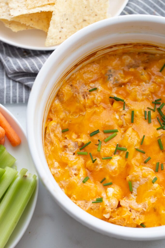 Overhead photo of a white casserole dish of buffalo chicken dip topped with green onions. On the side are white plates of baby carrots, celery, and homemade tortilla chips.