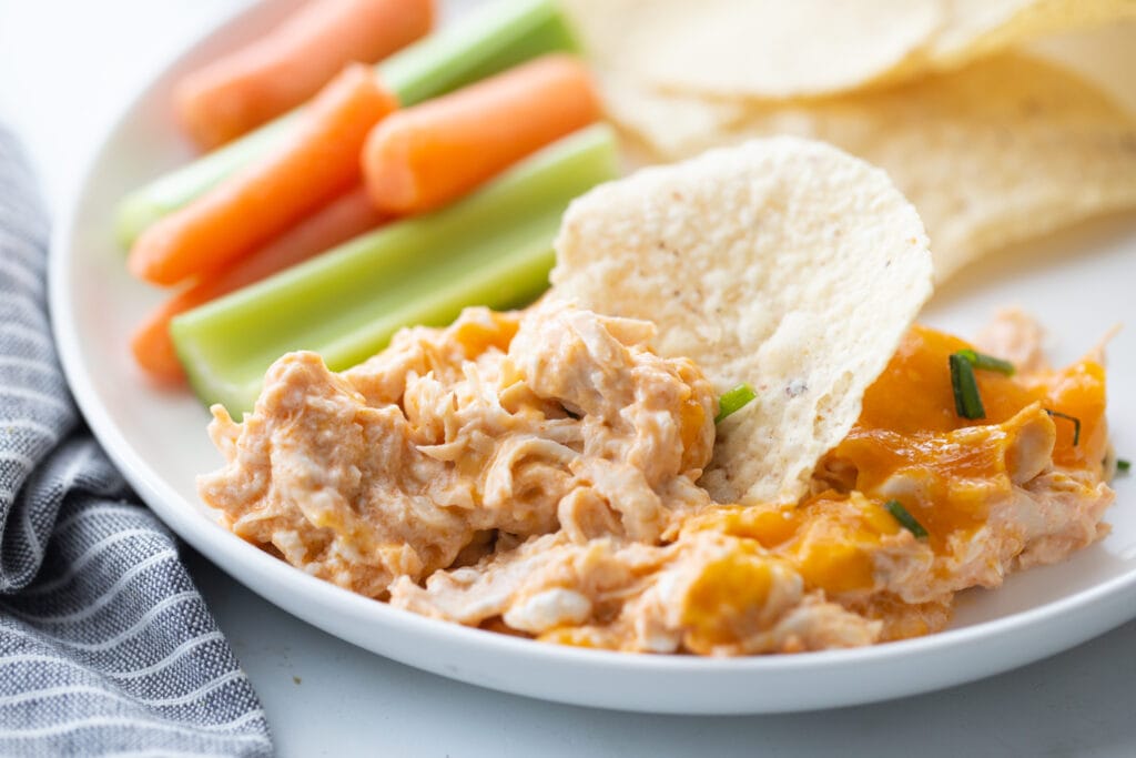 Hvit rimmed plate med buffalo kylling dip oppskrift og en chip stikker ut av toppen. Baby gulrøtter, selleri og tortilla chips er i bakgrunnen av platen.