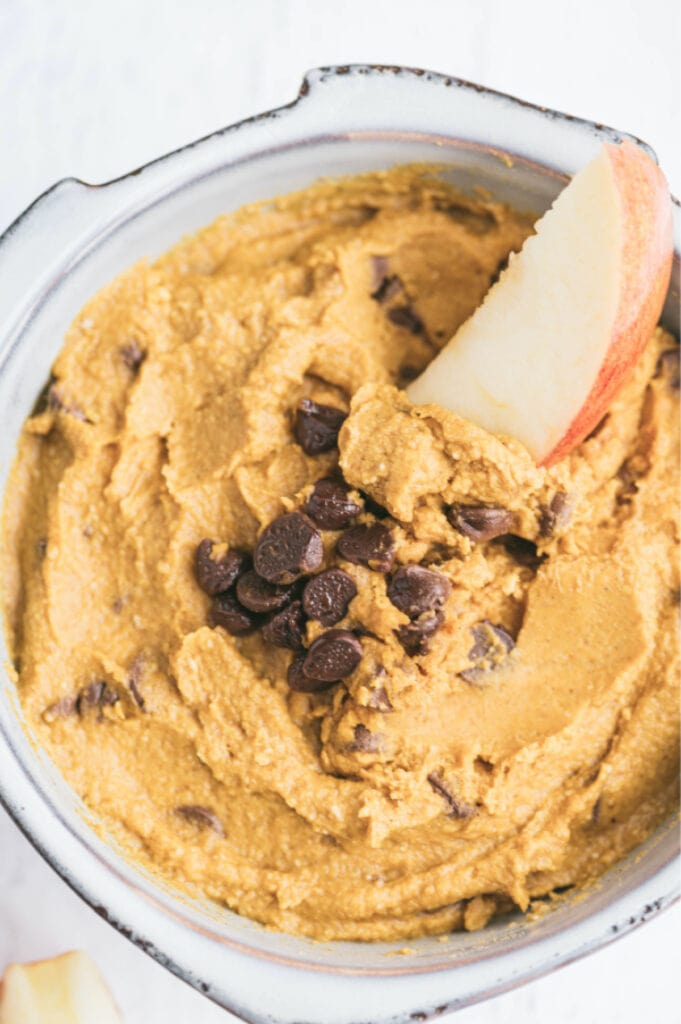 Overhead image of a white bowl of pumpkin pie hummus topped with chocolate chips, with an apple slice dipped in