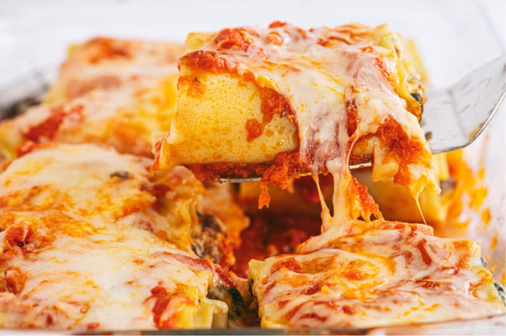 close up photo of a serving spatula lifting a lasagna roll out of a serving dish with gooey cheese trailing behind.