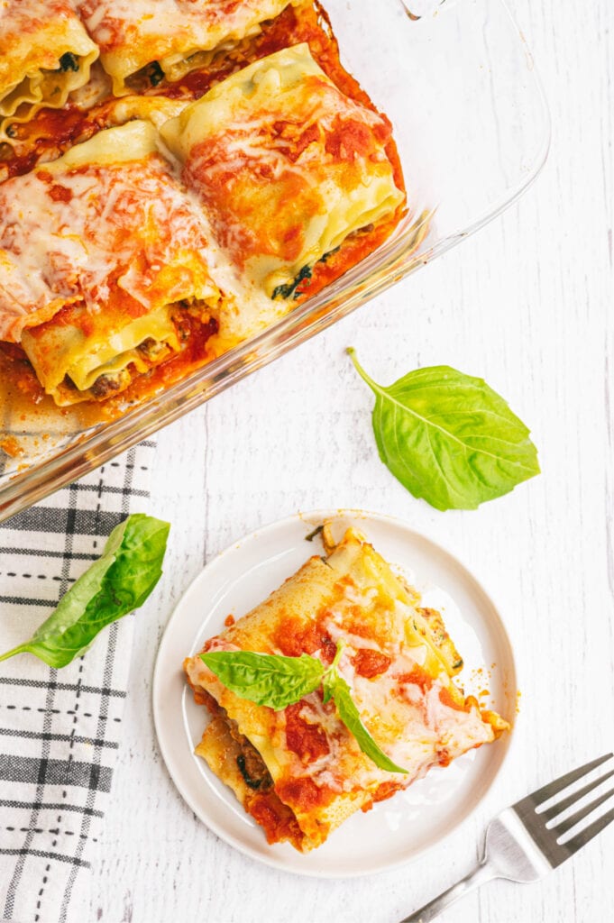 Overhead image: Glass baking dish of lasagna roll ups at top of image. At the bottom is a white rimmed plate with a single lasagna roll garnished with basil.