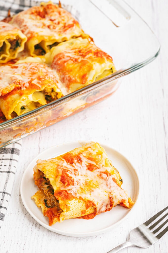 High angle photo of lasagna roll ups in a glass dish with a roll on a white plate and a fork nearby.