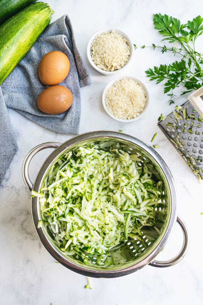 Overhead image: metal colander with shredded zucchini surrounded with zucchini fritter ingredients and a metal box grater.