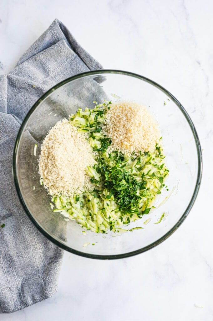 Overhead image: glass bowl of grated zucchini, bread crumbs, parmesan cheese, and herbs on a white background.