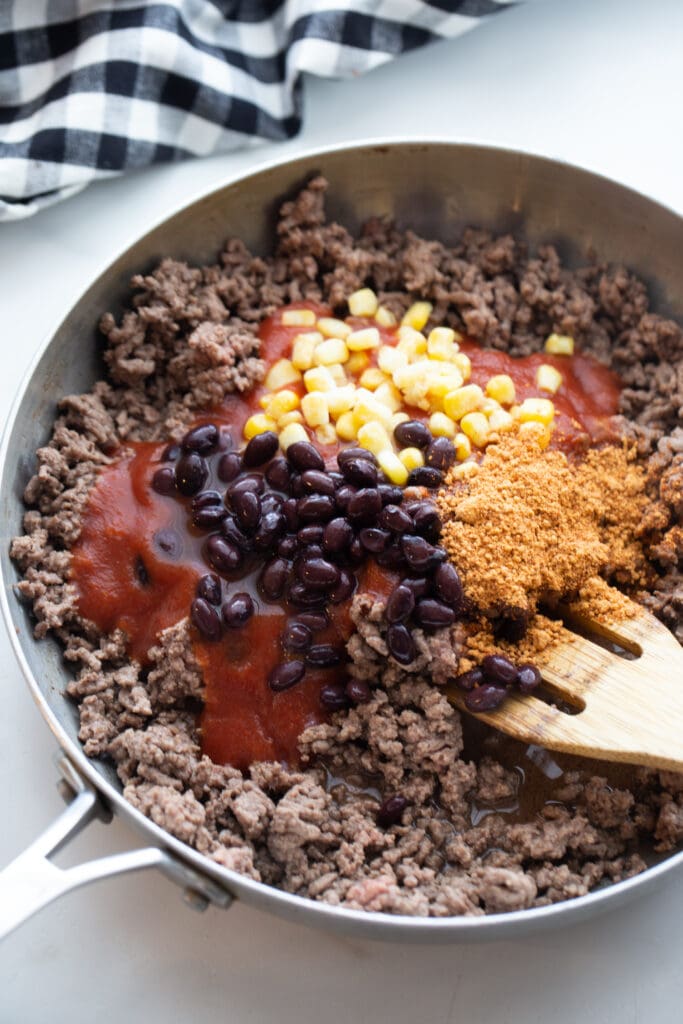 Overhead photo of cooked ground beef in a pan, layered with tomato sauce, corn, black beans, and spices, ready to be mixed up with a wooden spoon