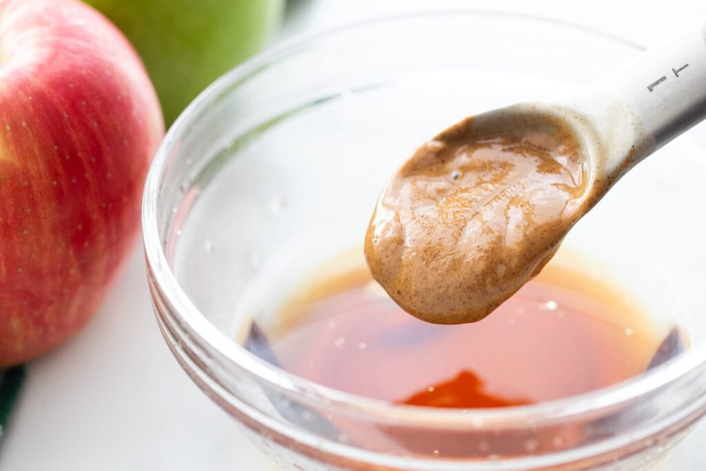 Almond butter on a measuring spoon being added to a small glass bowl of brown liquid.