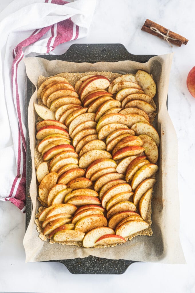 sliced apples sprinkled with cinnamon in rows on top of a crust on a parchment lined baking sheet, ready for the oven.