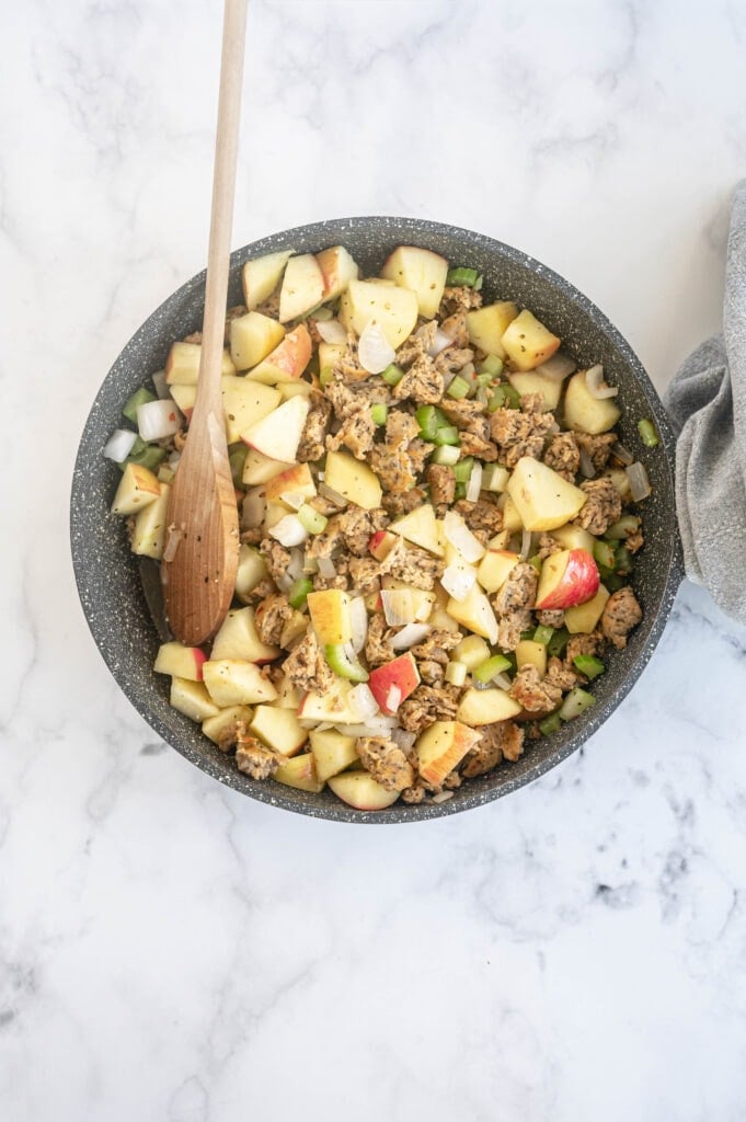 Overhead photo of poultry sausage, veggies, and apples in a skillet, with a wooden spoon resting on the side.