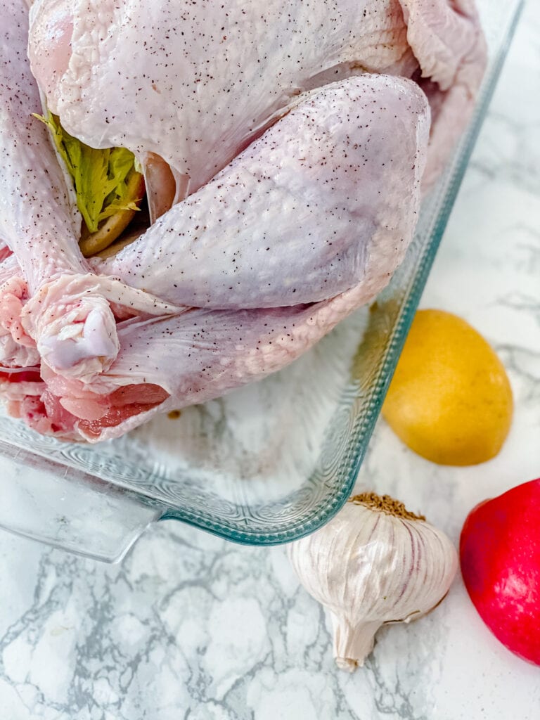 A whole raw turkey in a glass baking dish on a white marble surface. There is a whole lemon and head of garlic on the side.