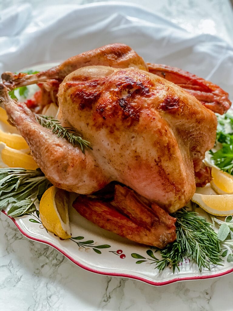 Whole turkey with crispy skin on a holiday plate and garnished with greens and lemon slices.