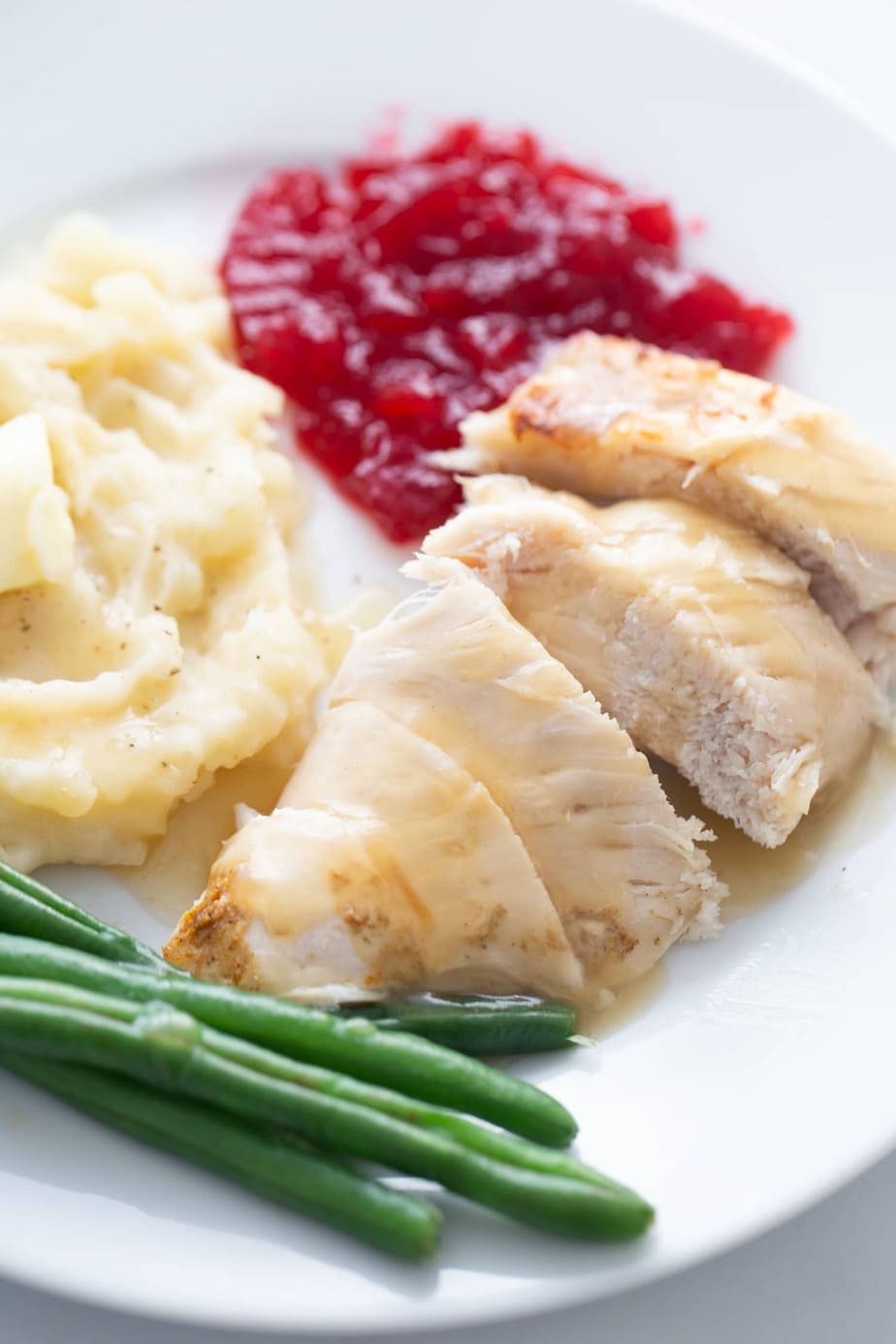 Close up photo of a white plate with sliced turkey breast, green beans, mashed potatoes, and cranberry sauce.