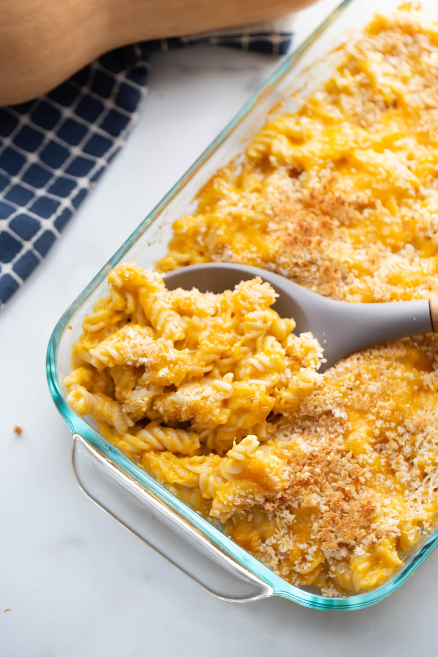 Glass baking dish of butternut squash mac and cheese topped with breadcrumbs with a gray serving spoon 