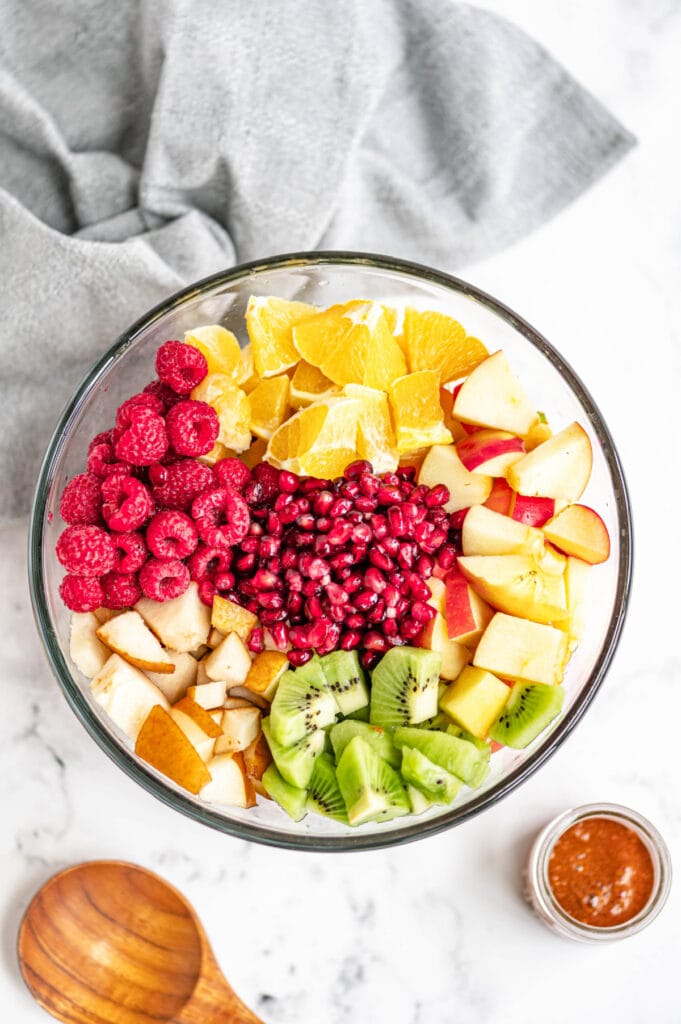 Fruits are diced and placed evenly in a large glass bowl on a white marble surface, near a wooden spoon and gray towel.