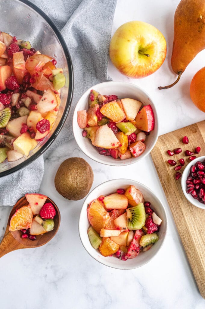 Two servings of fruit salad are placed in small white bowls, next to a wooden serving spoon.