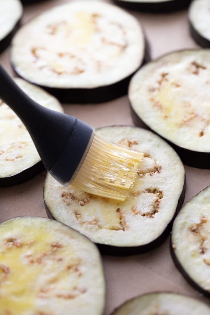 Olive oil is being brushed onto uncooked slices of eggplant for keto eggplant parmesan.